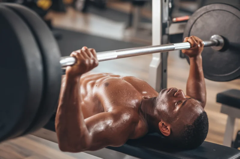 Shirtless man performing bench press with barbell in gym