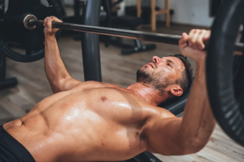 Shirtless man performing bench press with barbell in gym