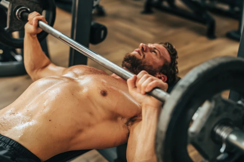 Shirtless muscular man performing bench press with barbell in gym