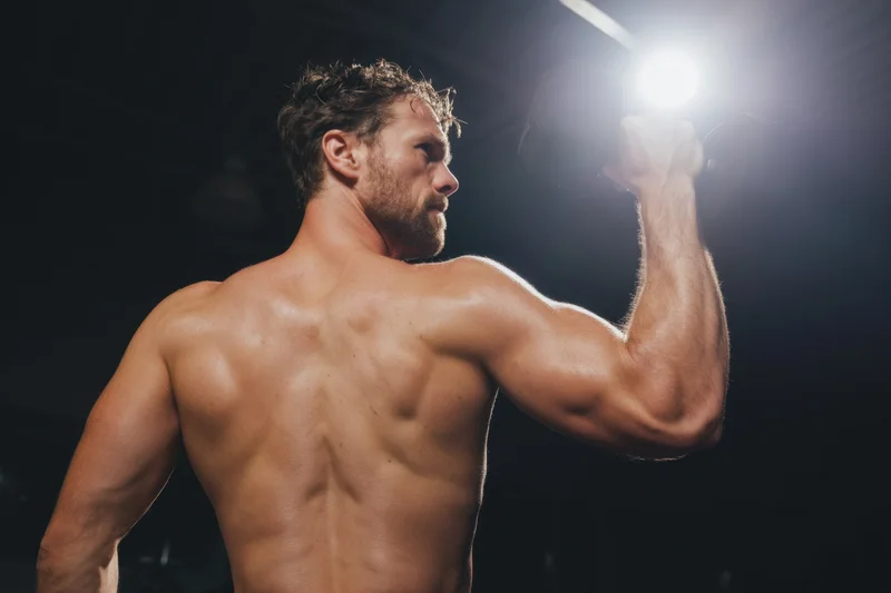 Shirtless muscular man lifting dumbbell in dim gym under spotlight