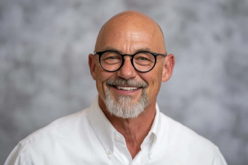 Bald man with gray goatee and black-rimmed glasses smiling in a white collared shirt against a neutral background