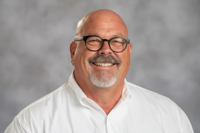 Bald man with gray goatee and black-rimmed glasses smiling in white shirt against gray background