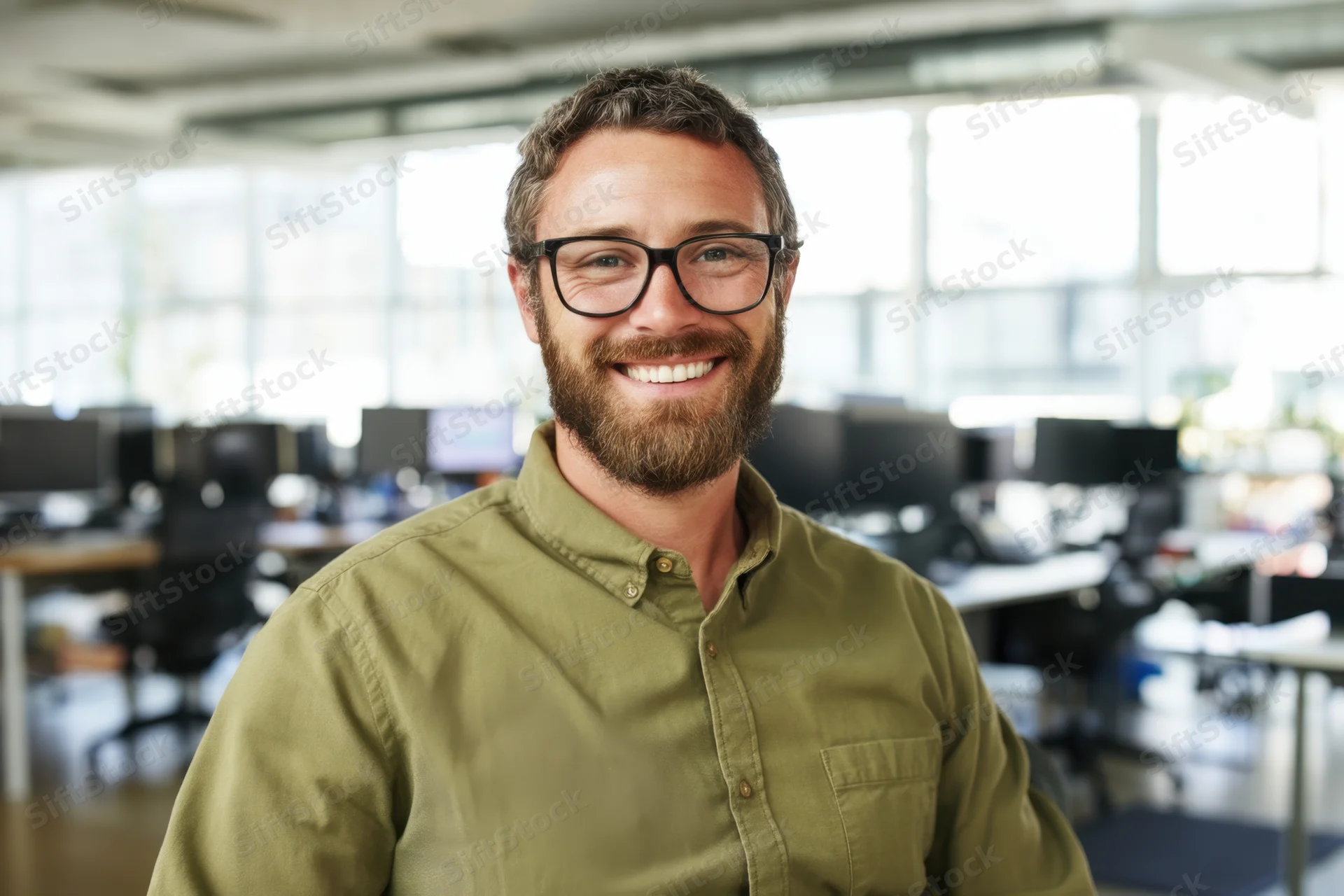 Smiling man in olive-green button-down shirt and glasses standing in modern office workspace
