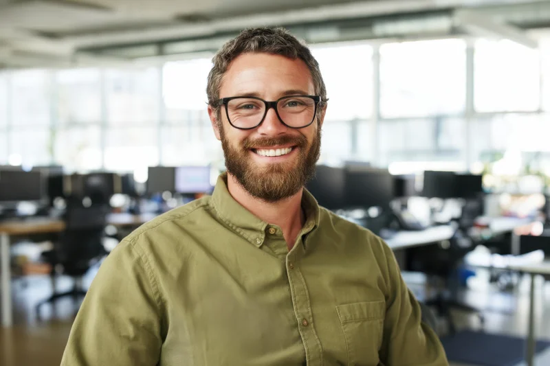 Smiling man in olive-green button-down shirt and glasses standing in modern office workspace