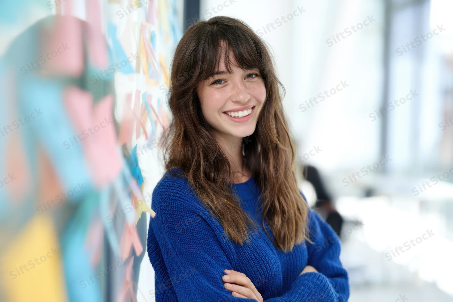 Smiling woman in blue sweater with arms crossed standing in office near whiteboard with colorful sticky notes