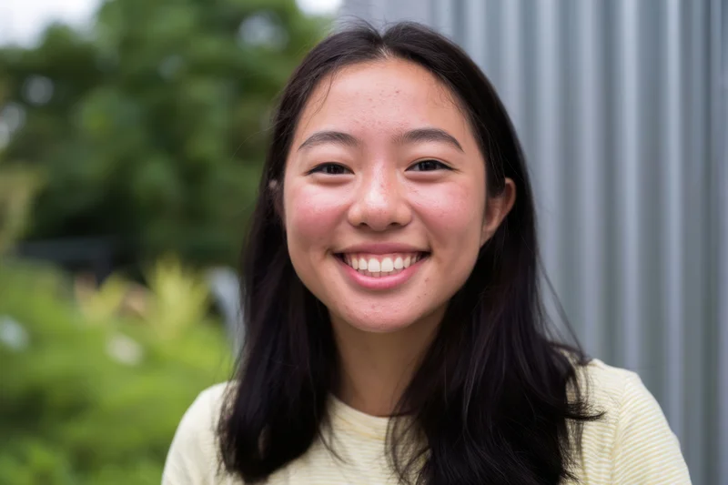 Young asian woman smiling at camera outdoors in front of green foliage and gray wall