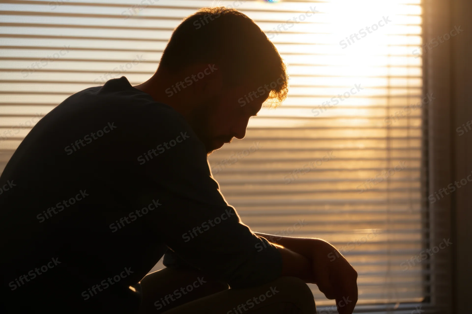 Man in silhouette sitting by window with head bowed and hand resting, backlit by warm sunlight through blinds