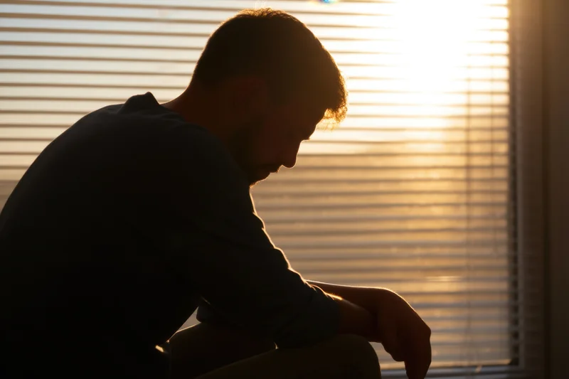 Man in silhouette sitting by window with head bowed and hand resting, backlit by warm sunlight through blinds