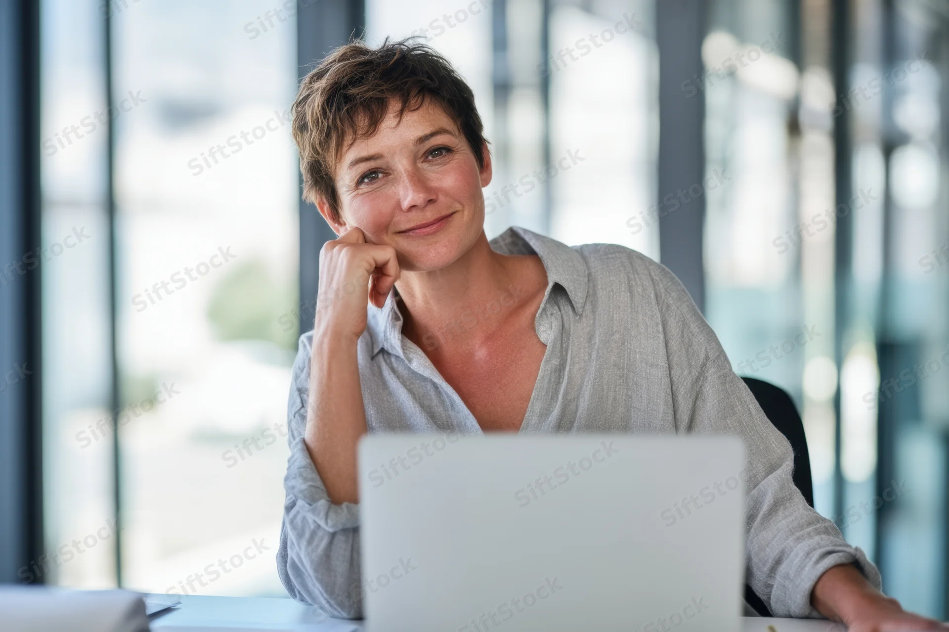 Woman sitting at desk with laptop in office, hand on chin, looking thoughtful