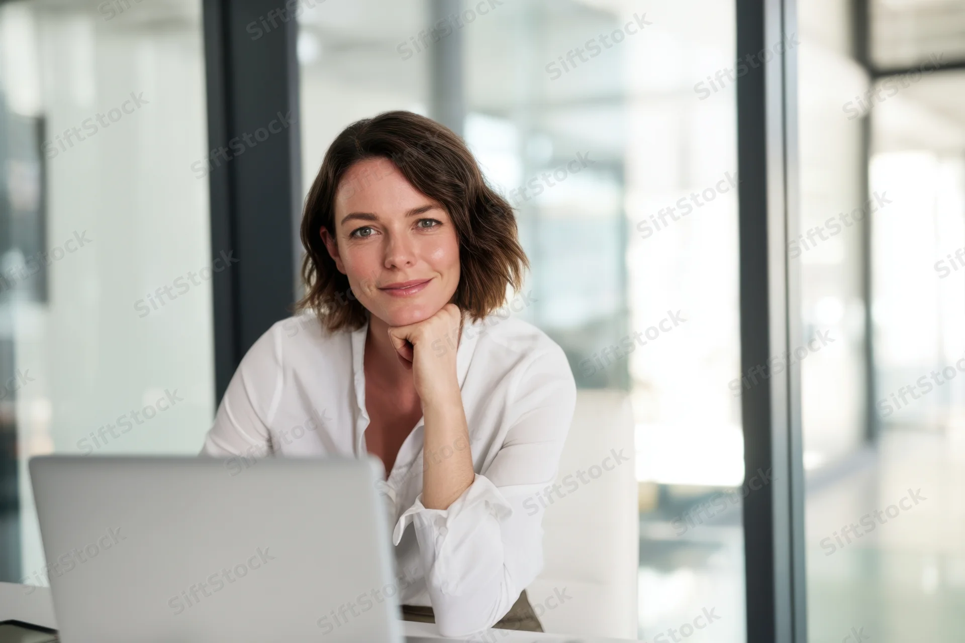 Woman sitting at desk with laptop in modern office, resting chin on hand, smiling thoughtfully
