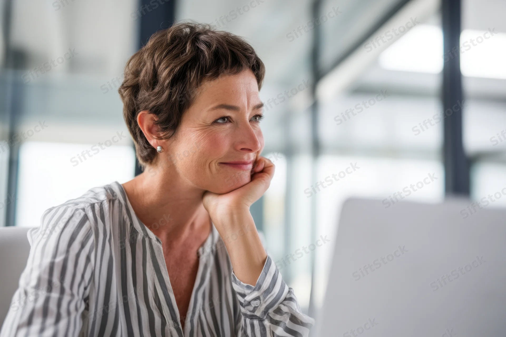 Woman smiling thoughtfully at desk in modern office with computer monitor and glass partitions