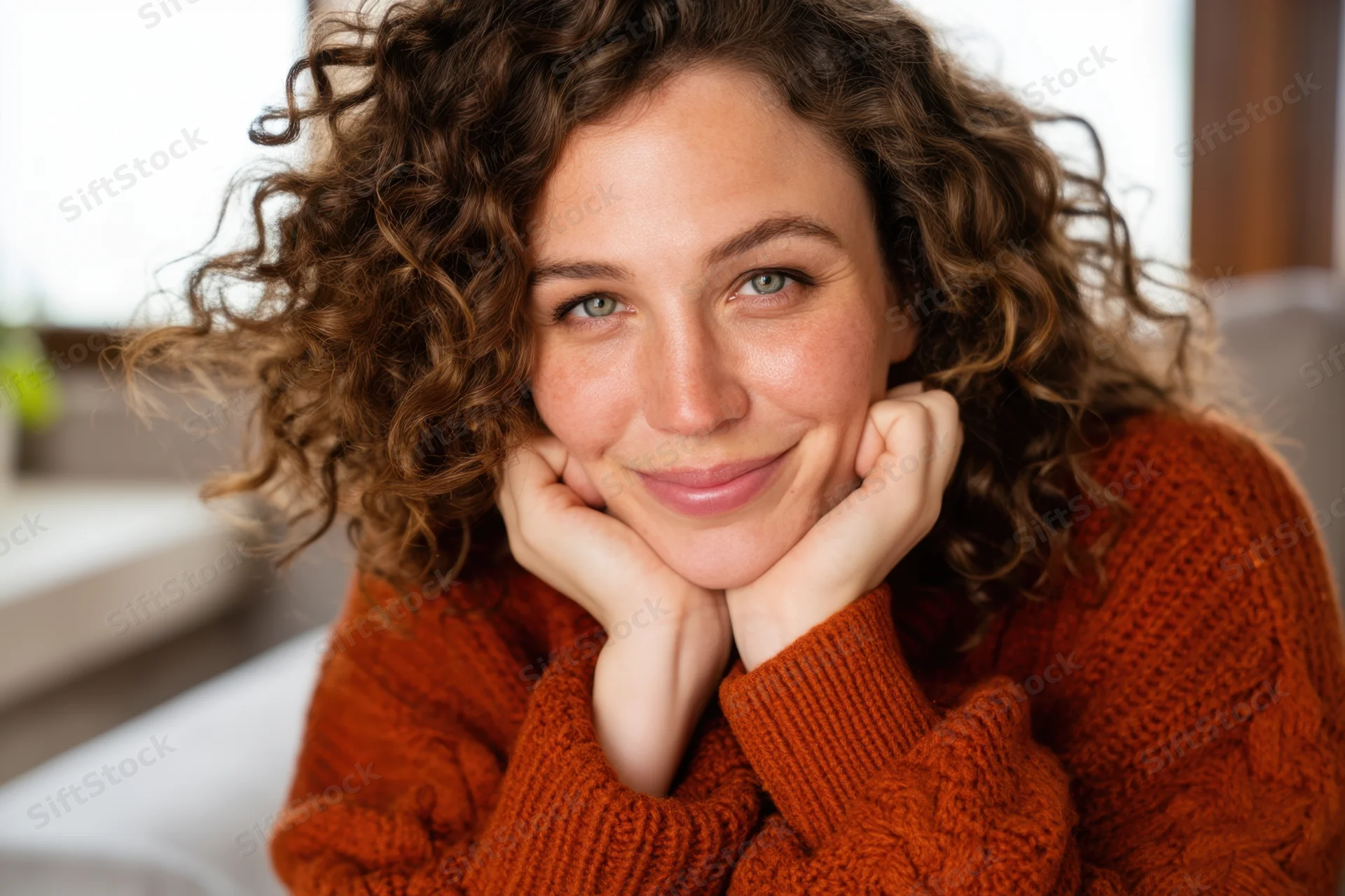 Woman with curly brown hair smiling while resting her chin on her hands indoors