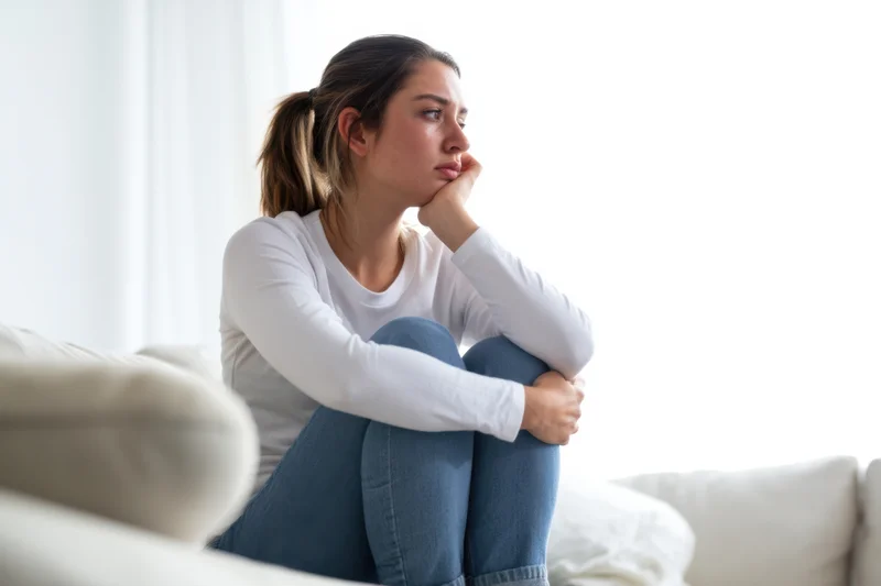 Young woman sitting on a light sofa in a bright living room, knees pulled up, hand on chin, looking away thoughtfully