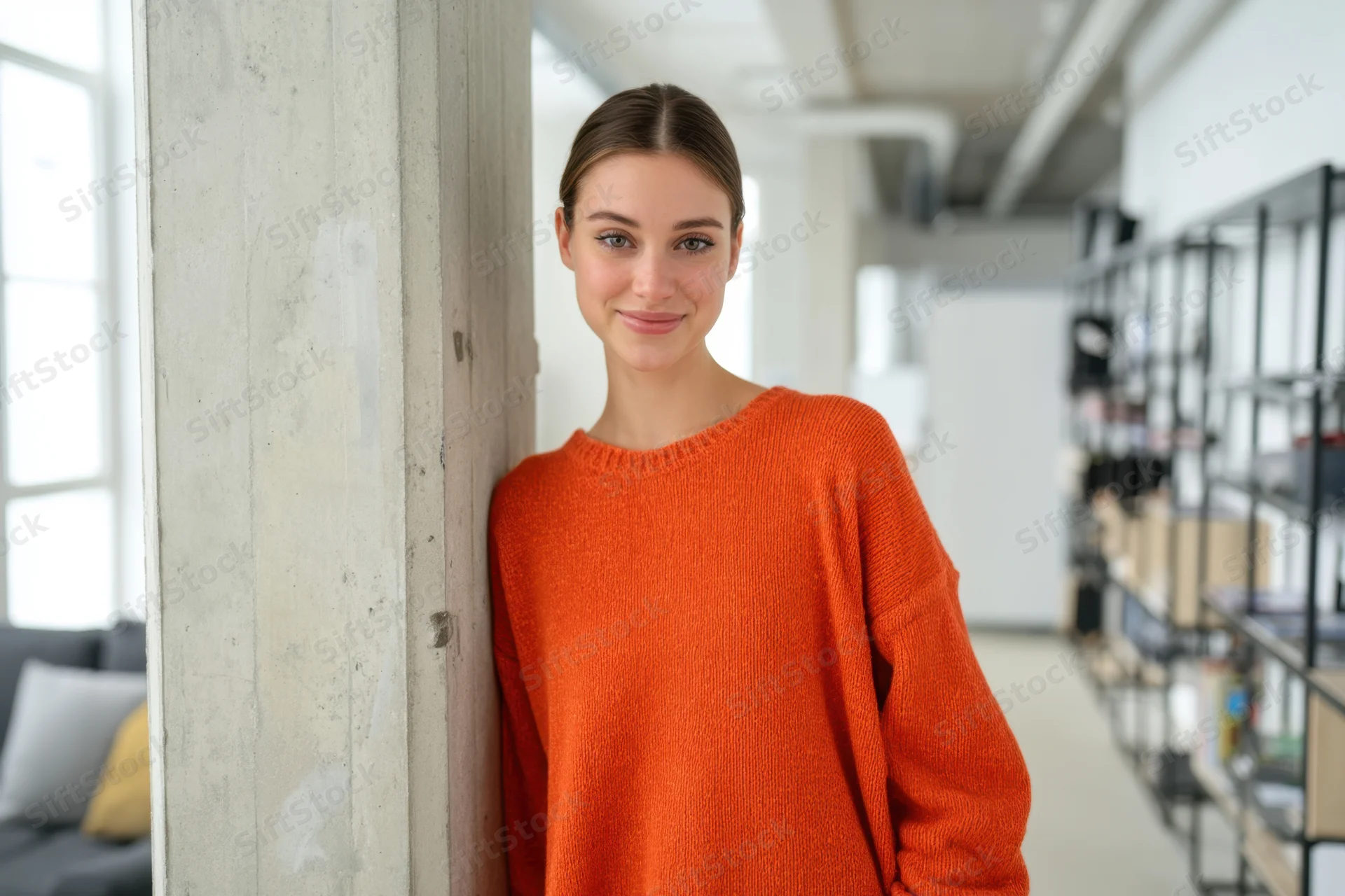 Woman in orange sweater leaning against concrete pillar in modern office space