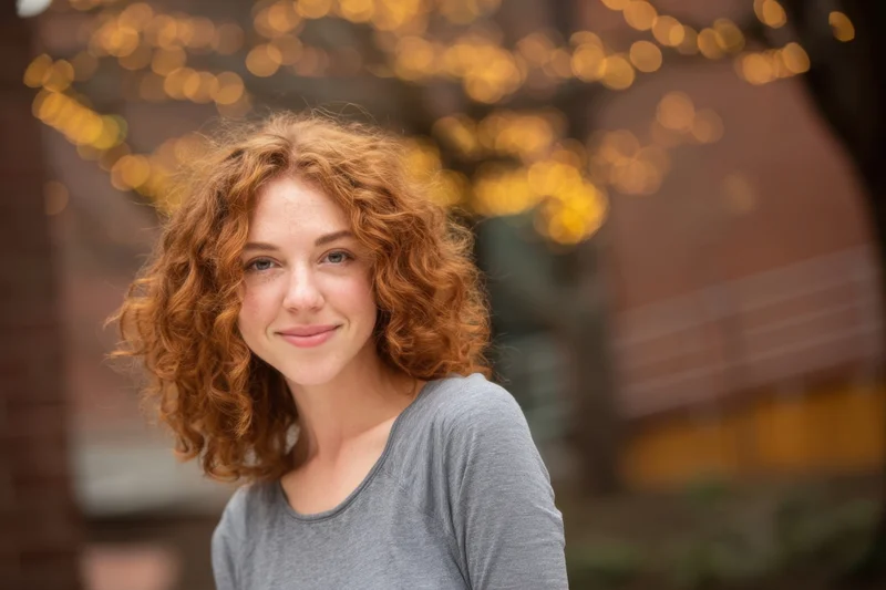 Woman with curly red hair smiling in outdoor city setting with warm bokeh lights behind her