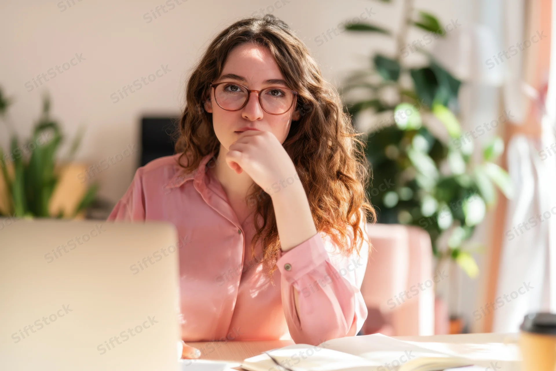 Woman with glasses thinking while working at a desk with a laptop in a sunlit room
