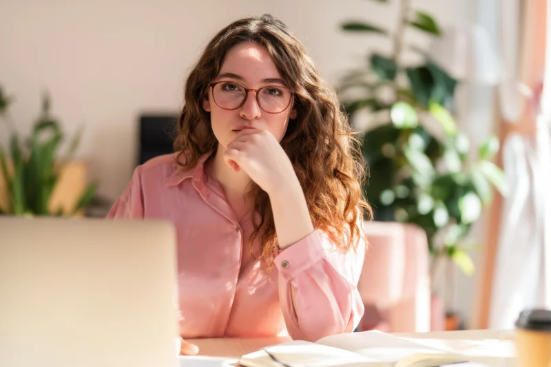 Woman with glasses thinking while working at a desk with a laptop in a sunlit room