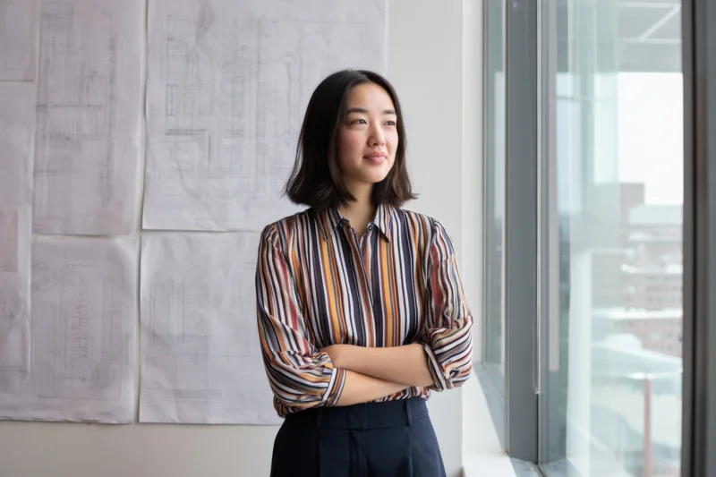 Woman architect standing by window in office with blueprints on wall