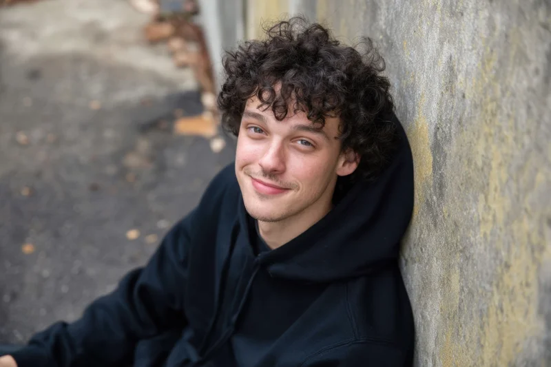 Young man with curly dark hair in black hoodie leaning against concrete wall smiling