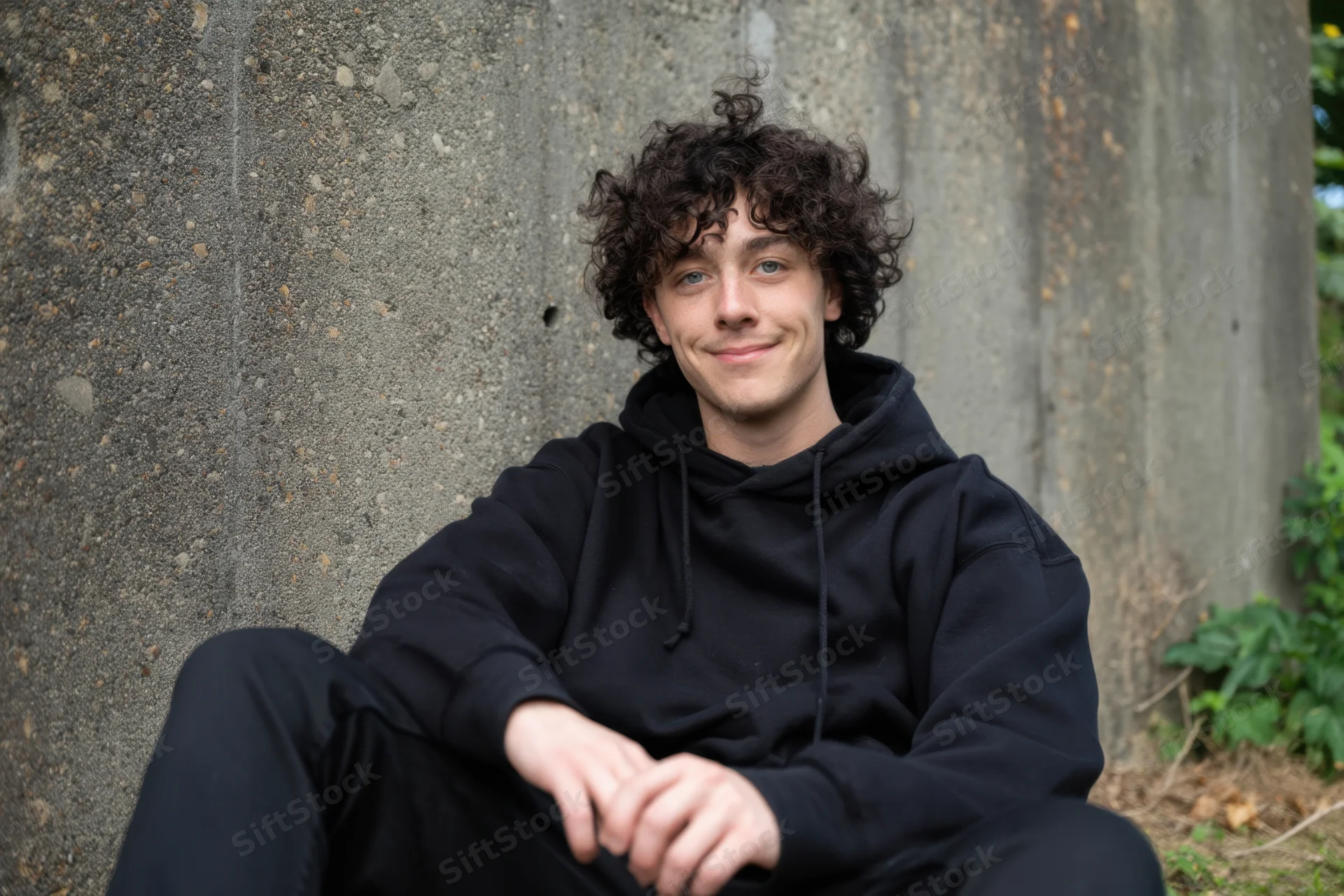 Young man with curly dark hair wearing black hoodie sitting against concrete wall outdoors smiling