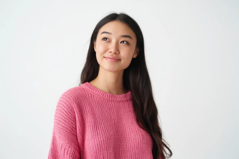 Young woman in pink knit sweater smiling slightly while looking to the side against white background