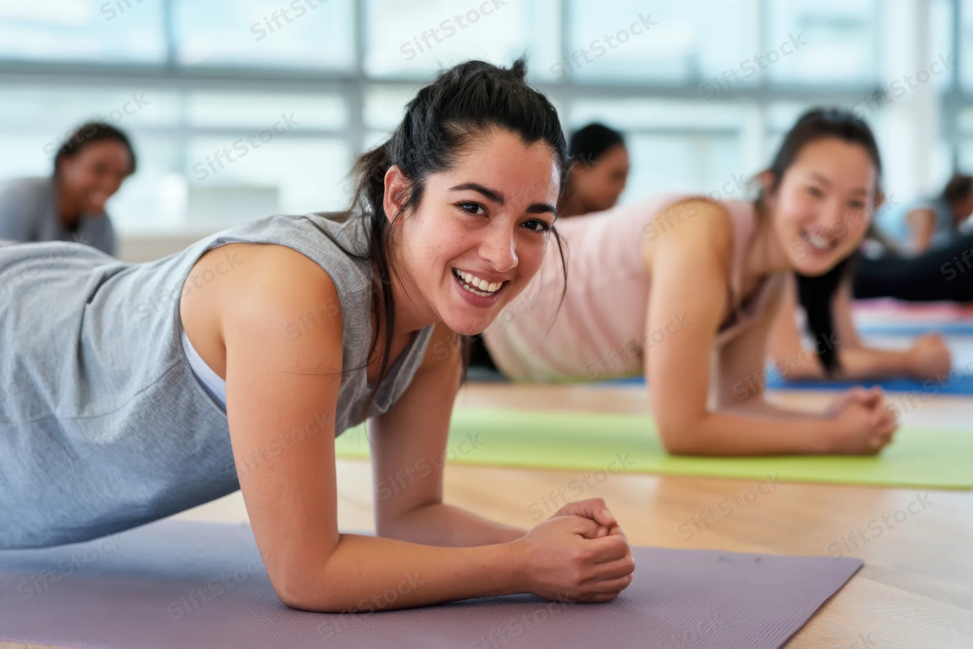 Woman smiling while holding plank position on yoga mat in bright modern studio with other women exercising in background