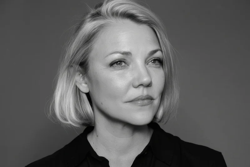 Grayscale portrait of a woman with short blonde hair and black shirt, looking directly at camera in studio setting