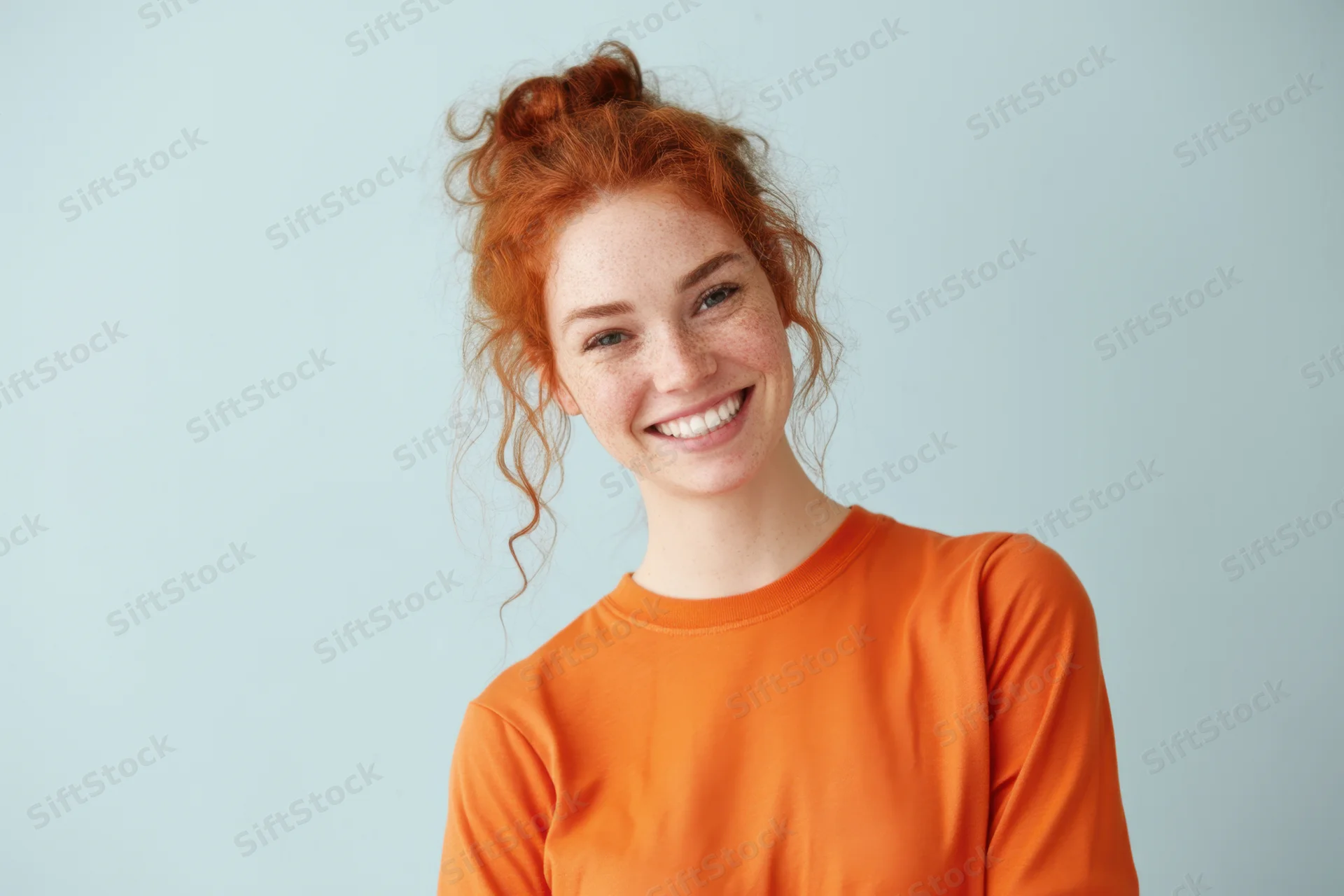 Smiling woman with red curly hair in a loose bun wearing an orange sweatshirt against a light blue background