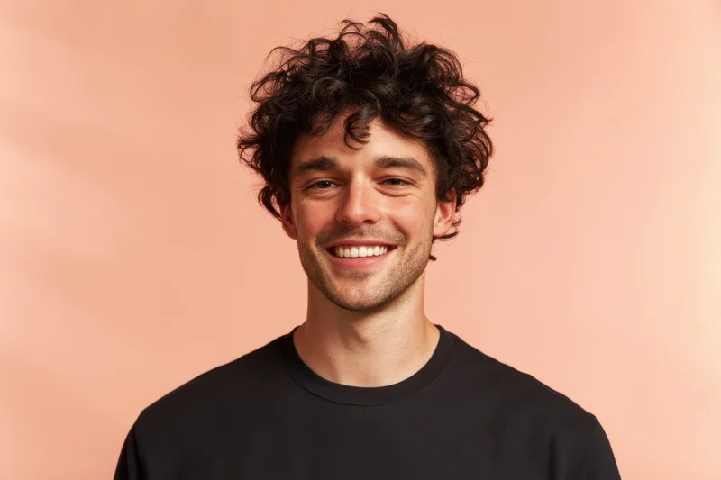 Young man with dark curly hair smiling in a black tshirt against a peach background