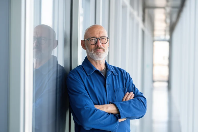 Bald man with glasses in blue jacket standing with arms crossed leaning against glass window in office hallway