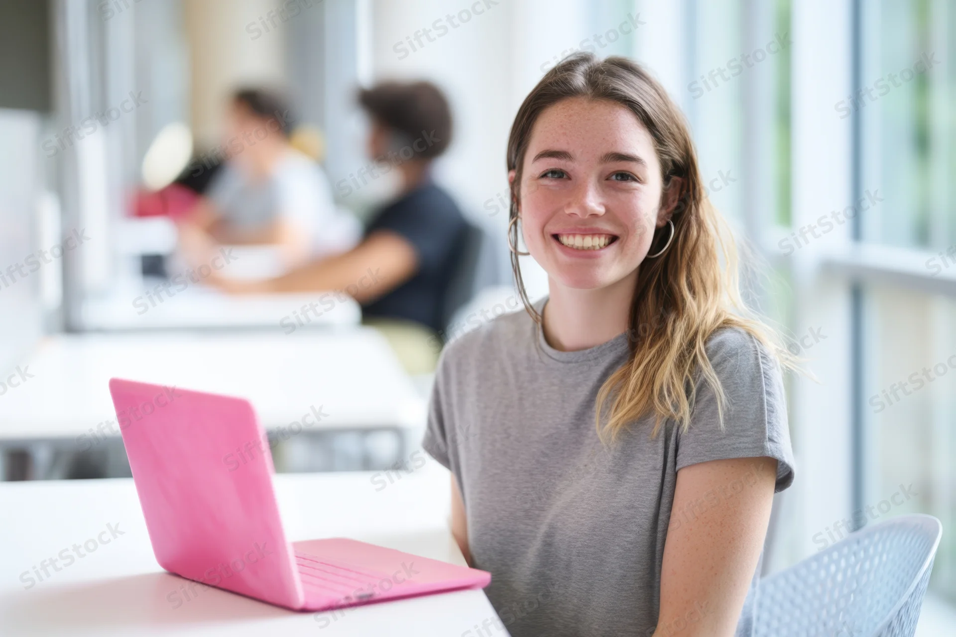 Young woman smiling at camera with pink laptop on desk in bright modern office workspace