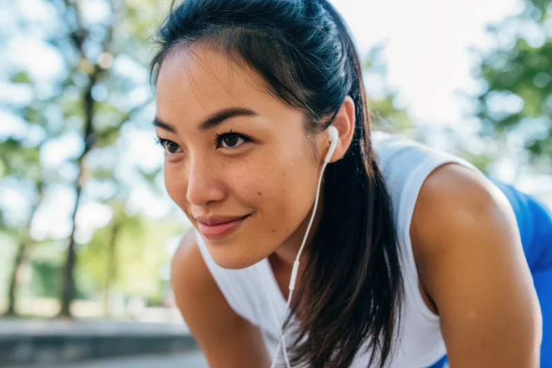 Fit woman smiling during outdoor workout in park wearing white athletic top and blue leggings with earphones