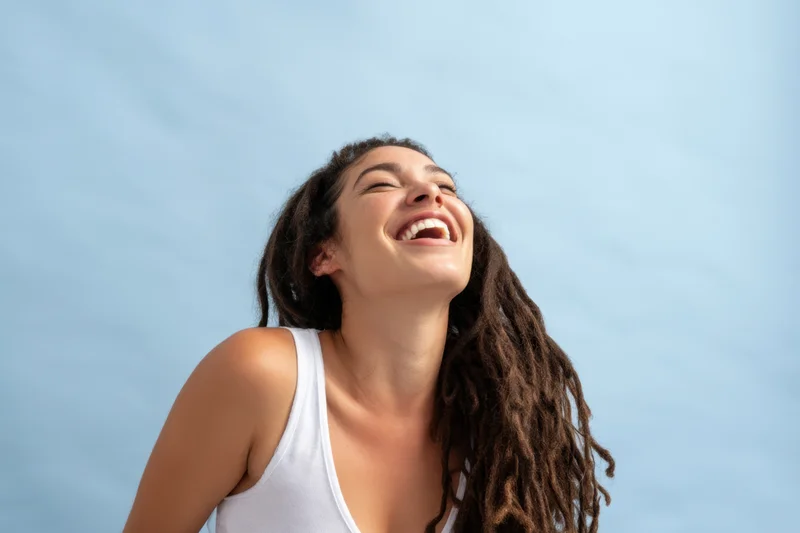 Woman laughing with head tilted back against blue sky wearing white tank top