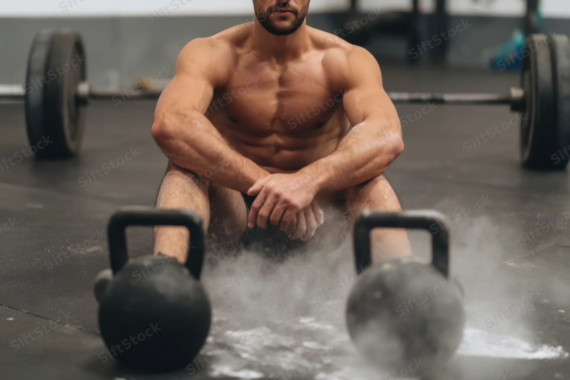 Muscular man sitting between kettlebells in a gym, dust rising from the floor around him