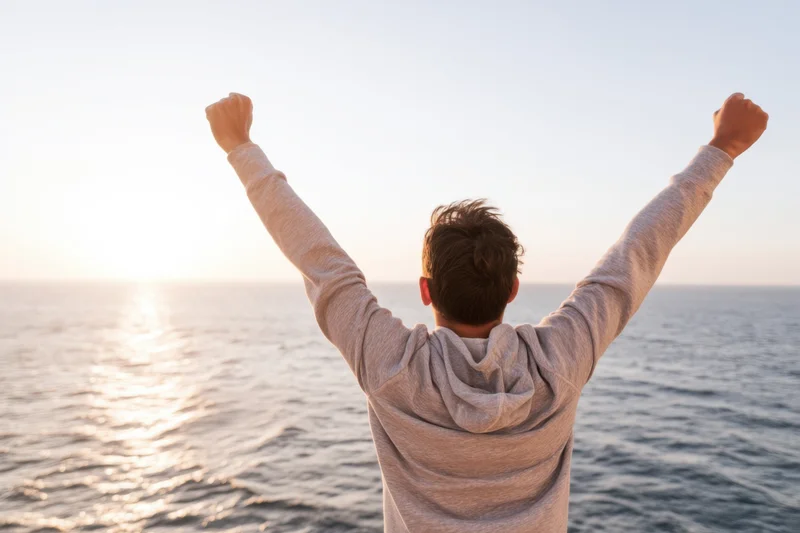 Man with arms raised in victory facing ocean at sunrise