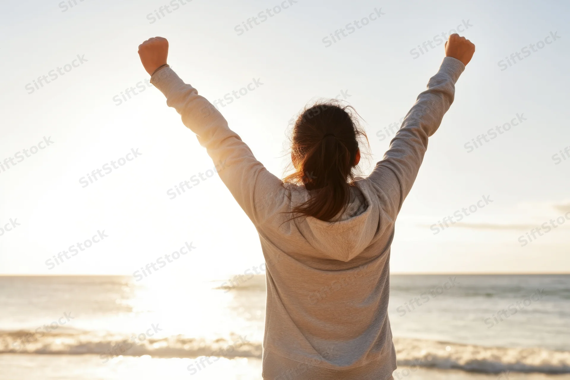 Woman with arms raised in victory facing ocean at sunset