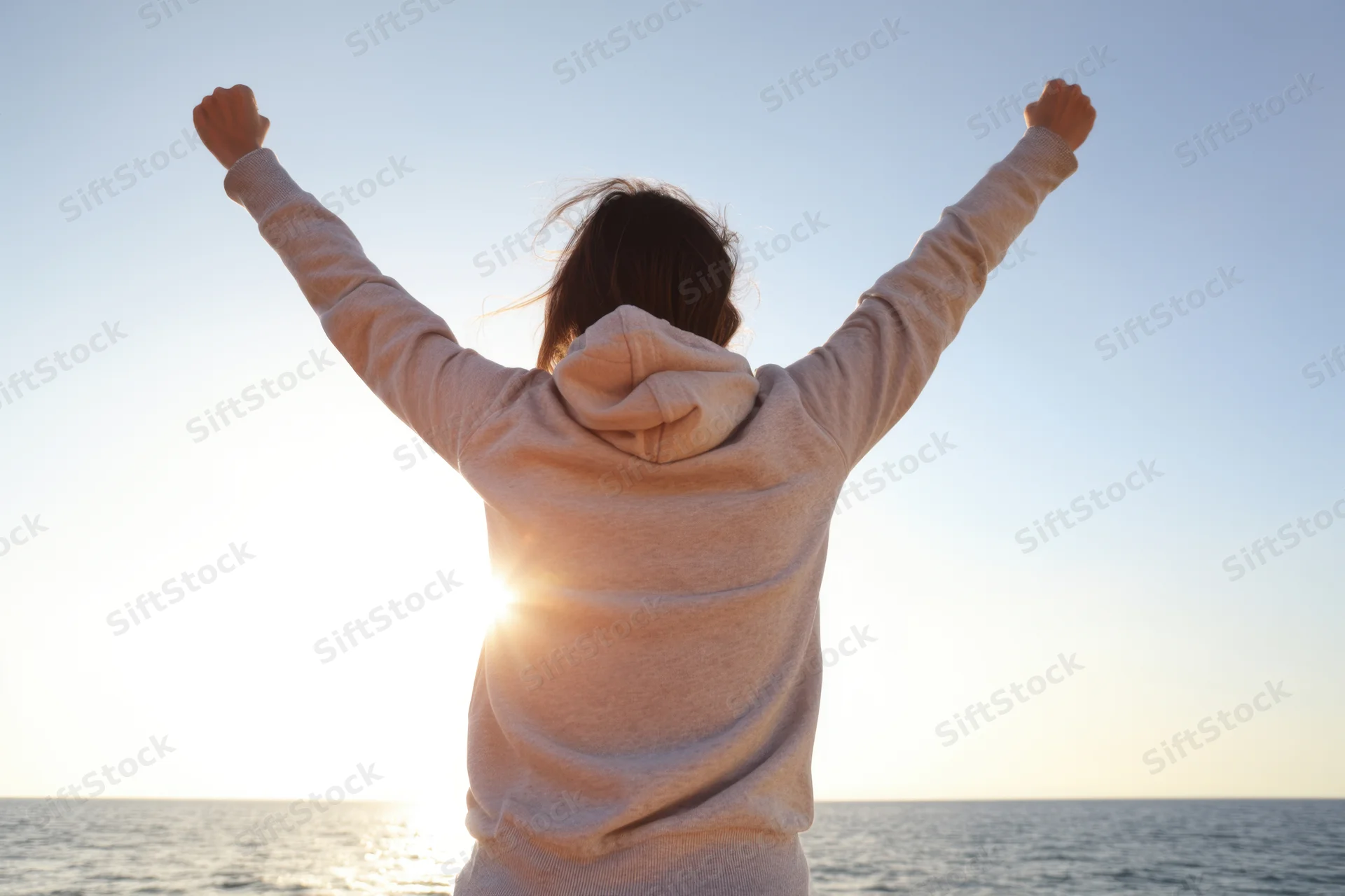 Person in a light hoodie with arms raised in victory at sunset