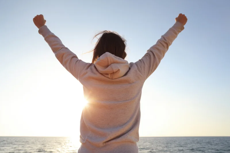Woman with arms raised in victory facing sunset over ocean