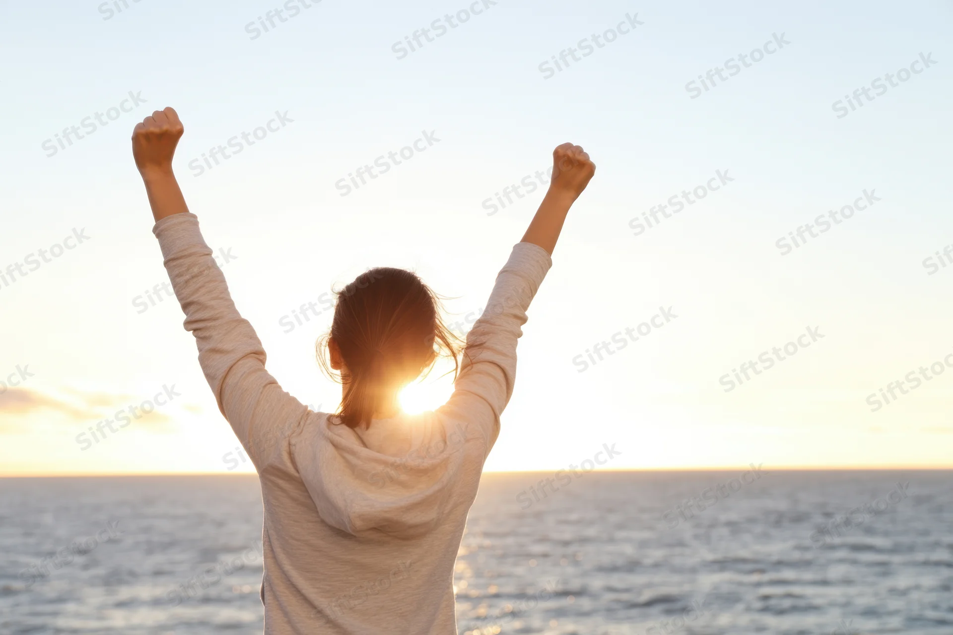Woman with arms raised in victory facing ocean at sunset
