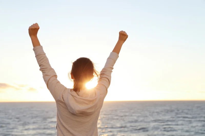 Woman with arms raised in victory facing ocean at sunset