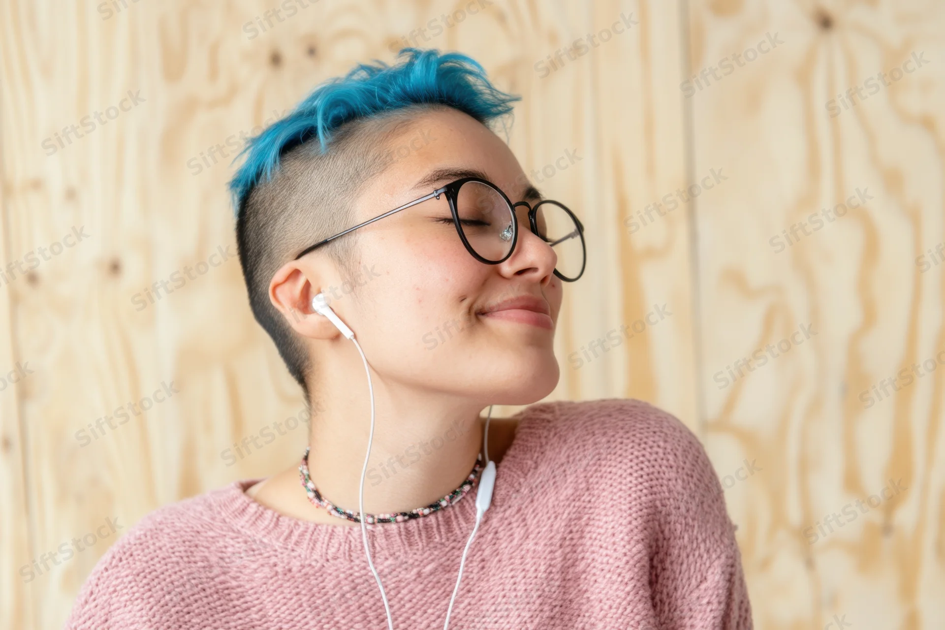 Woman with blue dyed short hair shaved on sides wearing round glasses and white earphones smiling with eyes closed against wooden background