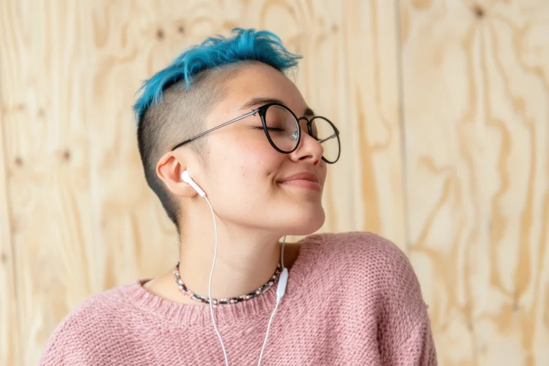Woman with blue dyed short hair shaved on sides wearing round glasses and white earphones smiling with eyes closed against wooden background