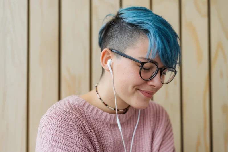 Woman with blue dyed short hair and undercut wearing glasses and white earphones, smiling, in pink sweater against wooden background