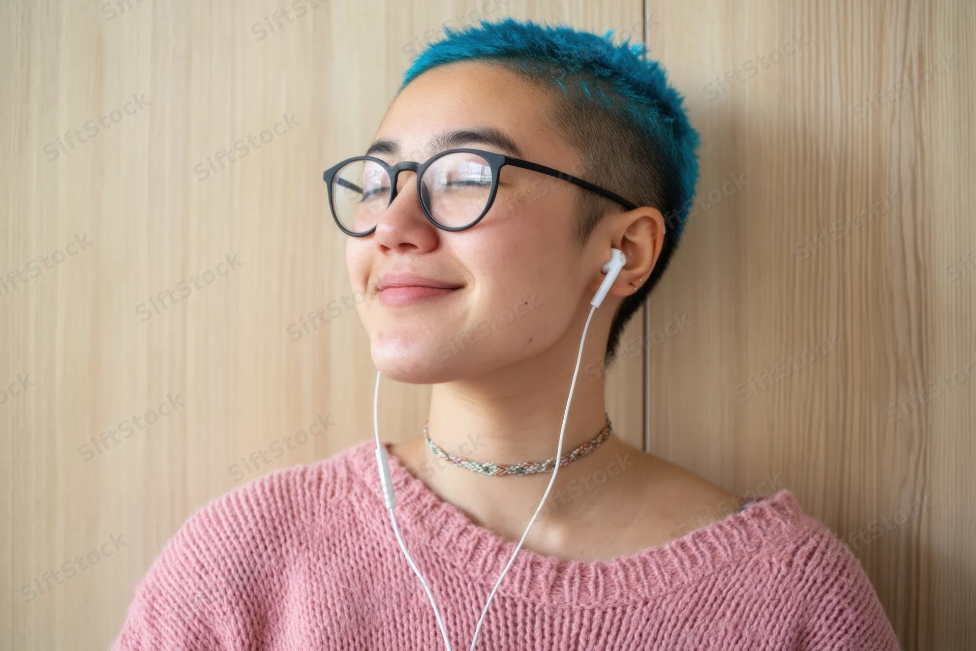 Woman with short blue-dyed hair shaved on sides wearing glasses and white wired earphones, smiling while leaning against wooden wall in pink sweater