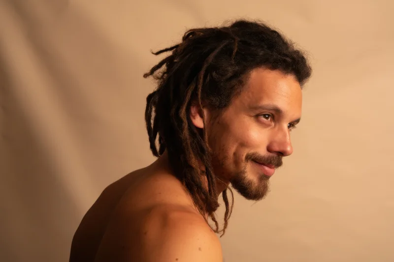 Shirtless man with medium-length dreadlocks in profile view smiling slightly against a warm beige studio background
