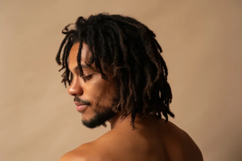 Shirtless man with medium-length dreadlocks in profile view against neutral background