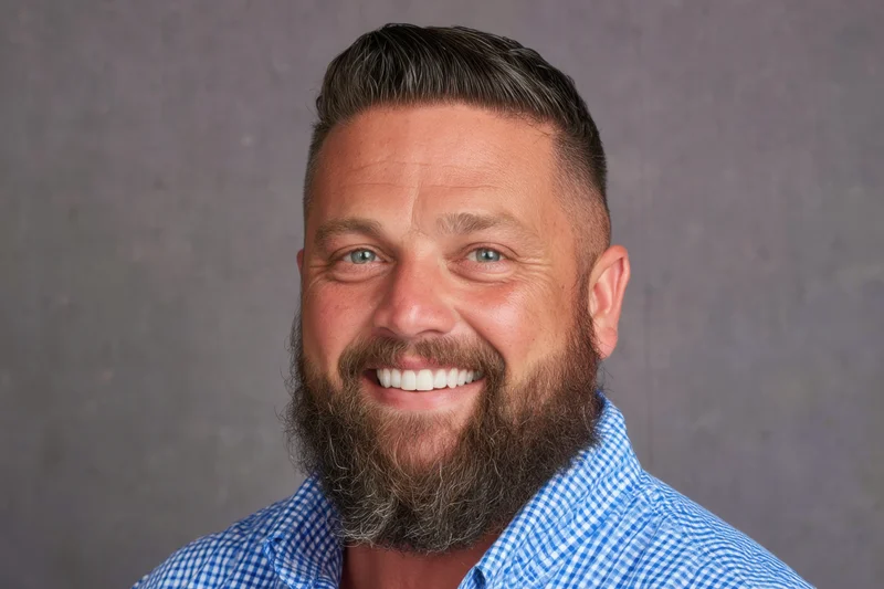 Smiling man with full beard and styled hair in blue checkered shirt against gray background