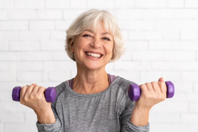 Smiling senior woman holding purple dumbbells in front of white brick wall