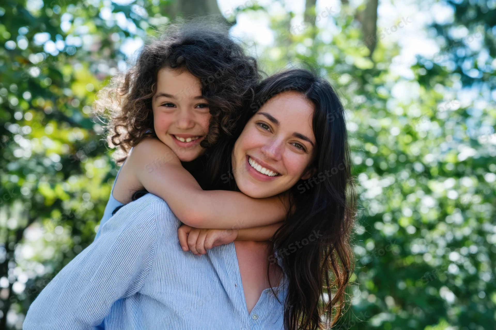 Woman carrying child on her back, both smiling, in a sunlit park surrounded by green trees