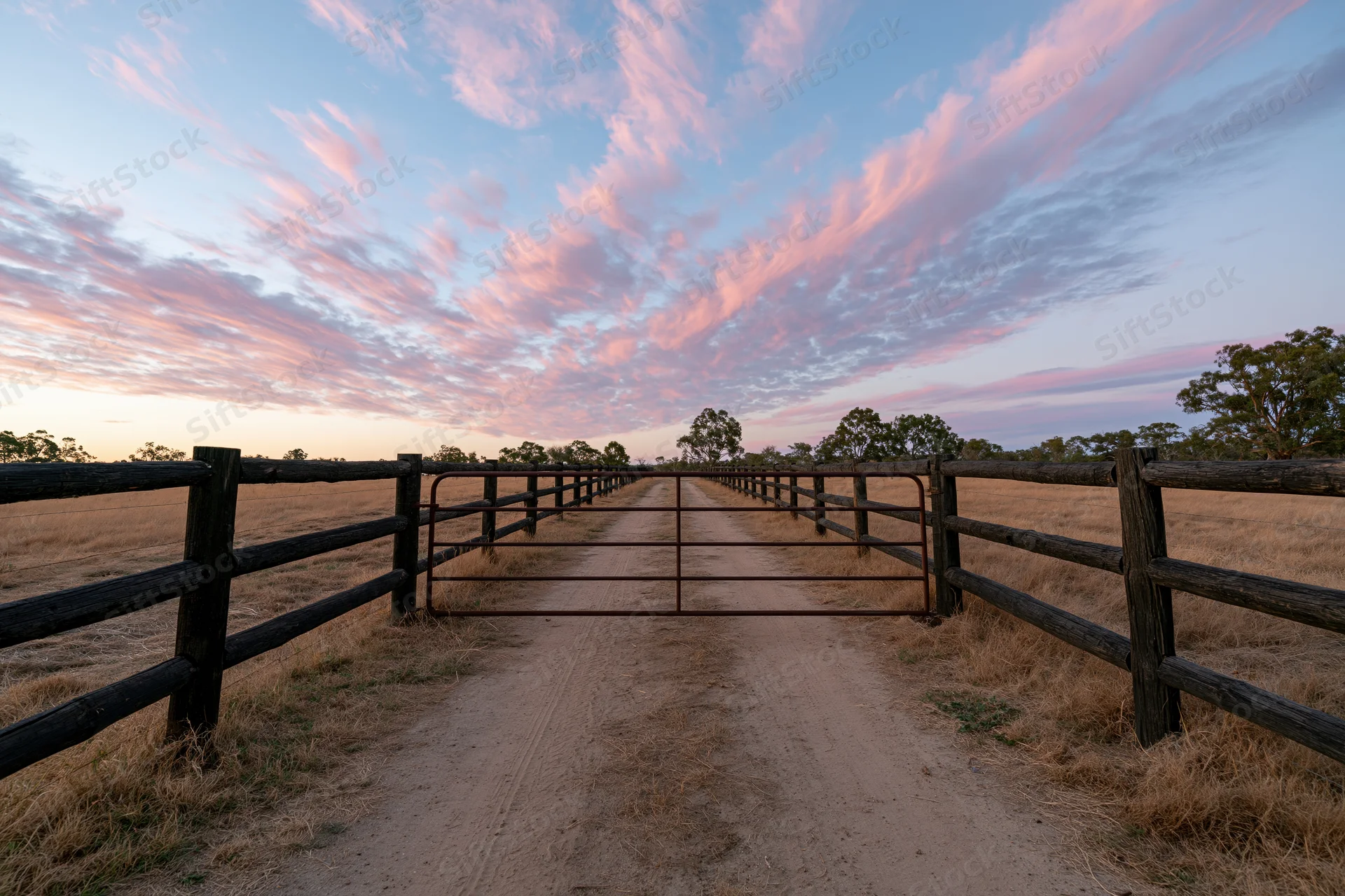 Metal farm gate flanked by dark timber post-and-rail fences along a dirt path at sunset